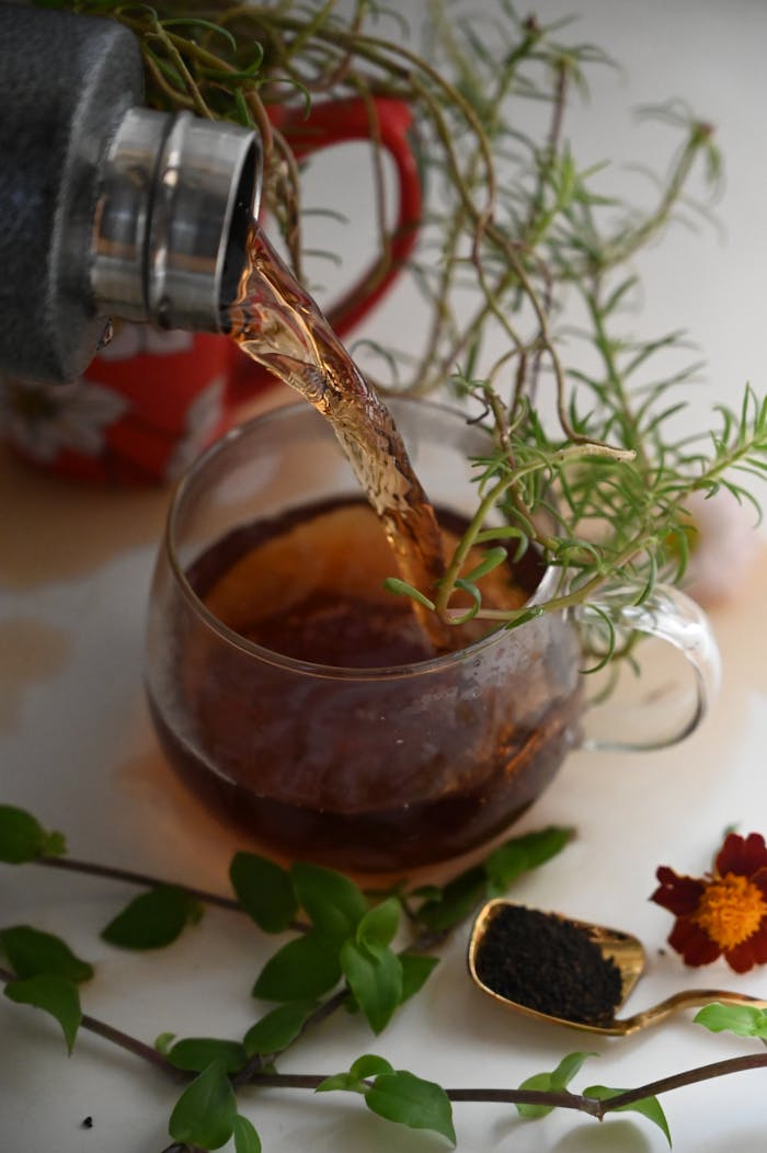 A cozy scene of herbal tea being poured into a glass cup surrounded by fresh greenery and flowers.
