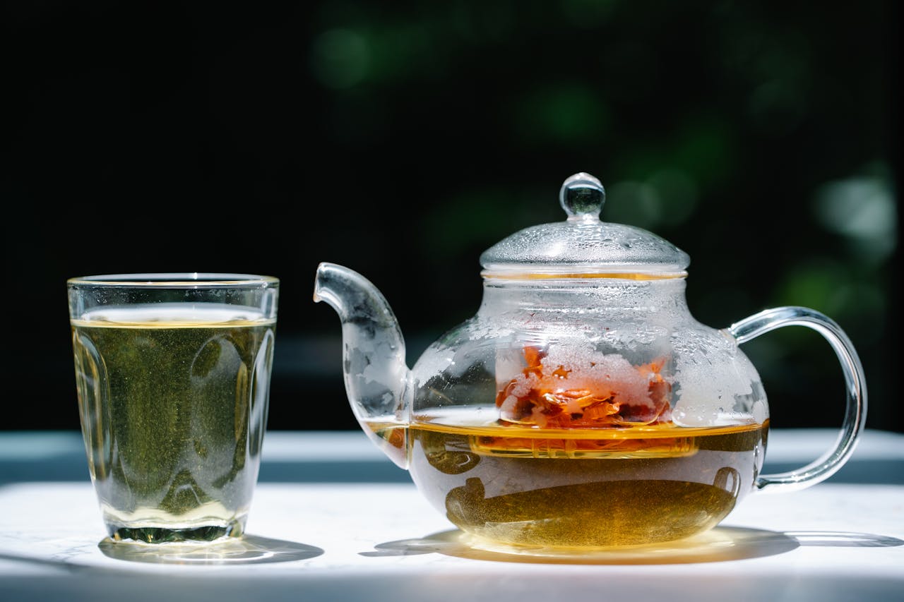 A glass teapot filled with herbal tea next to a transparent glass under sunlight. Refreshing and aromatic.